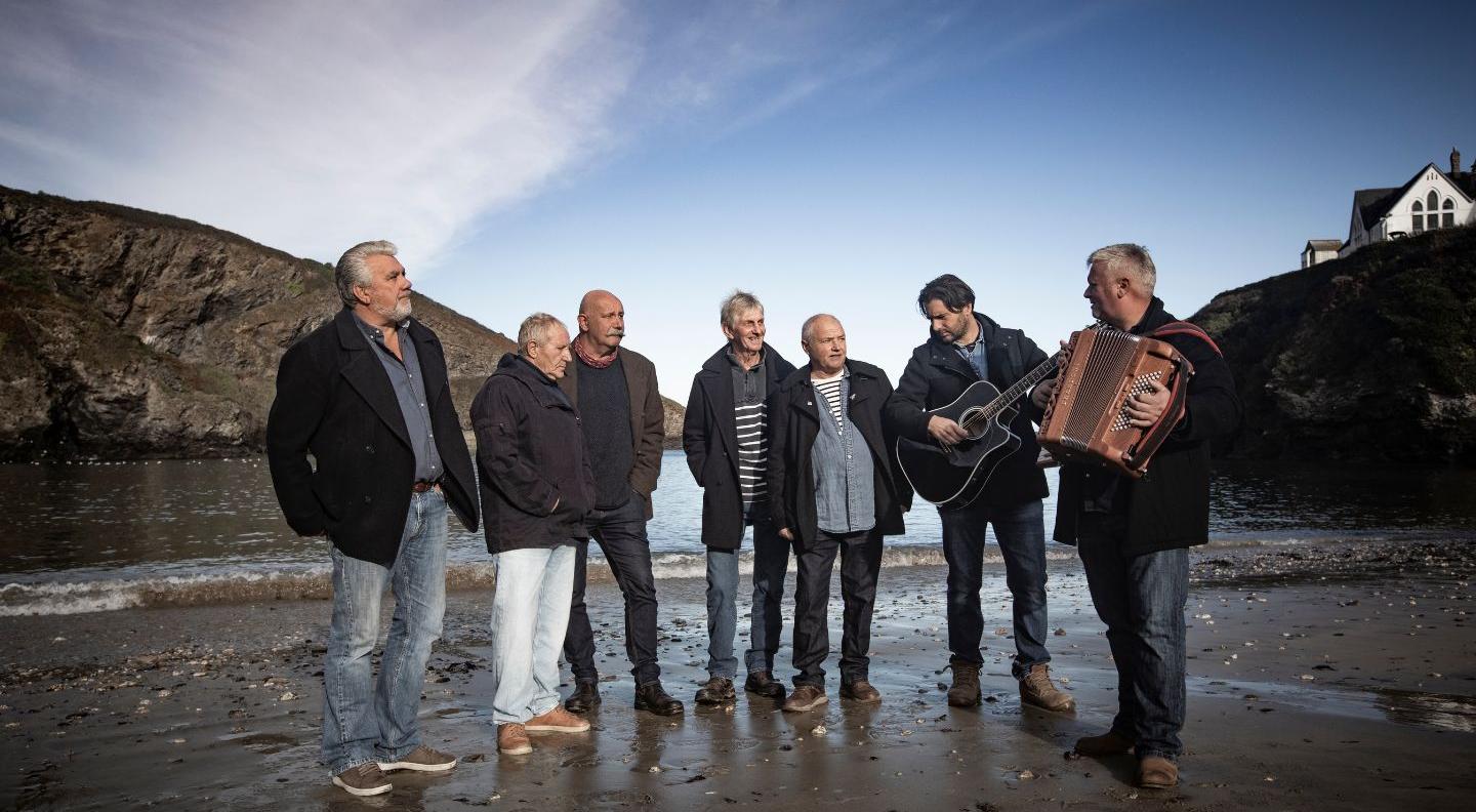 Seven men wearing jeans, jackets and boots stand on a sandy beach. There are cliffs on either side, and the sky is blue. One man is playing a guitar, and another a wooden accordion