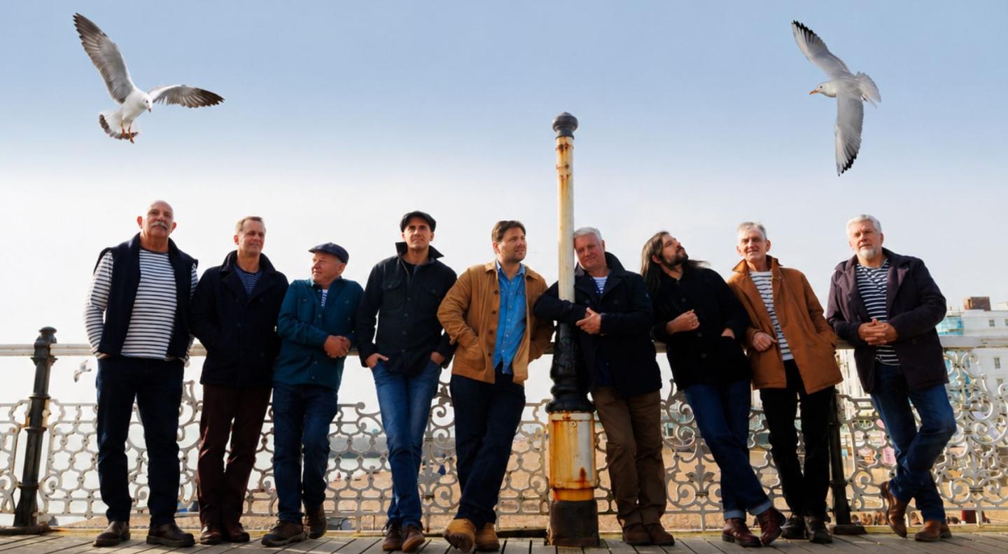 The nine members of Fisherman's Friends standing on a pier with seagulls flying above