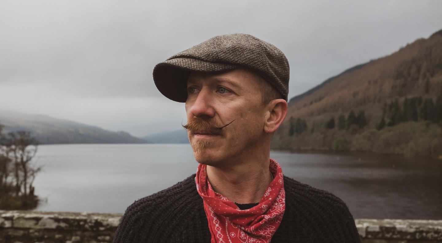 Foy Vance stands in front of a low stone wall with a loch or lake and hills behind him. The sky is grey.He wears a tweed flat cap, a dark top and a red bandana around his neck. He has a goatee and an impressive handlebar moustache which has been twisted into fine points at either end 