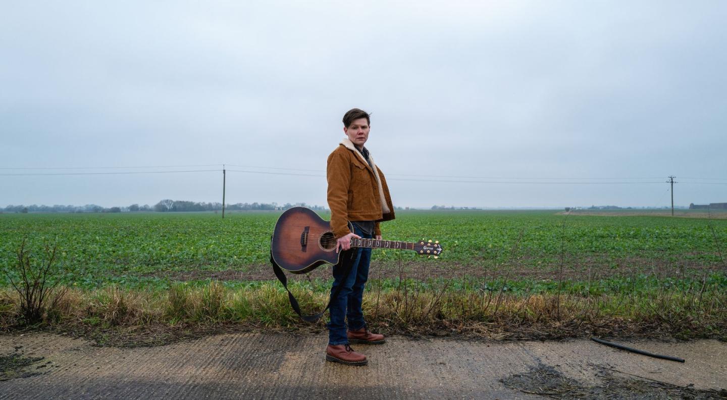 Grace Petrie standing in front of an open field, holding her guitar in one hand