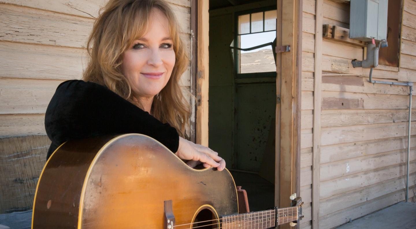 Gretchen Peters is seated with a guitar on her lap, in front of a wooden building