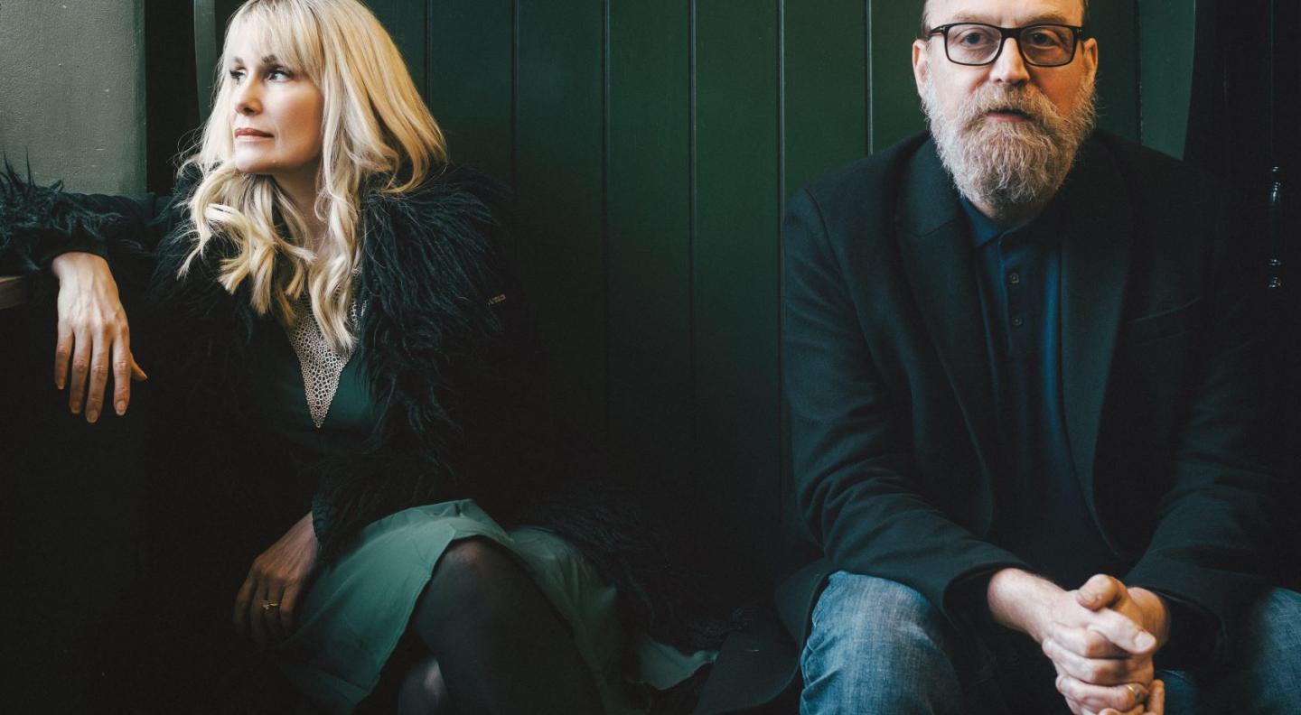 A white woman with long blonde hair and a white man with a beard and glasses sit side by side. He stares at the camera, she leans off looking to the left