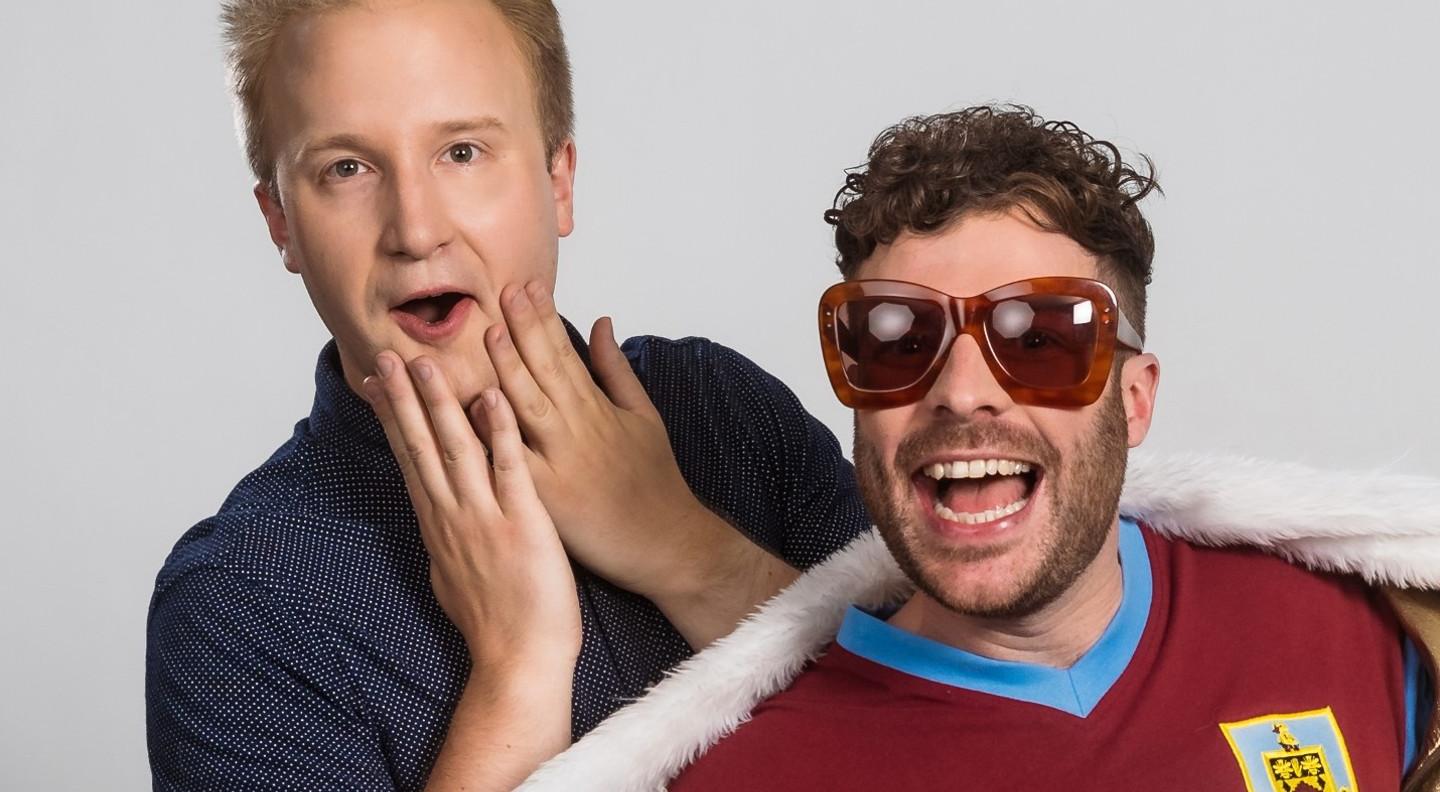 William Hanson stands behind Jordan North looking amused and shocked with his hands up to his open mouth. Jordan is slightly crouched in front of him as if he's just jumped into the shot. He has a large grin on his face and wears a Burnley Football Club top, a fake fur collar around his shoulders and oversized sunglasses