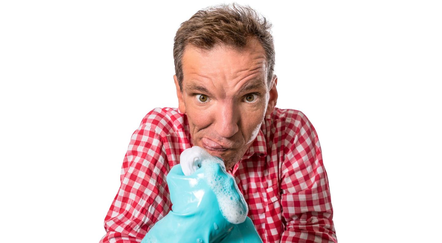 A full colour mid-shot of Henning Wehn looking at the camera with his tongue sticking out of the side of his mouth. He wears blue washing up gloves covered in soap suds and a red and white checked shirt.