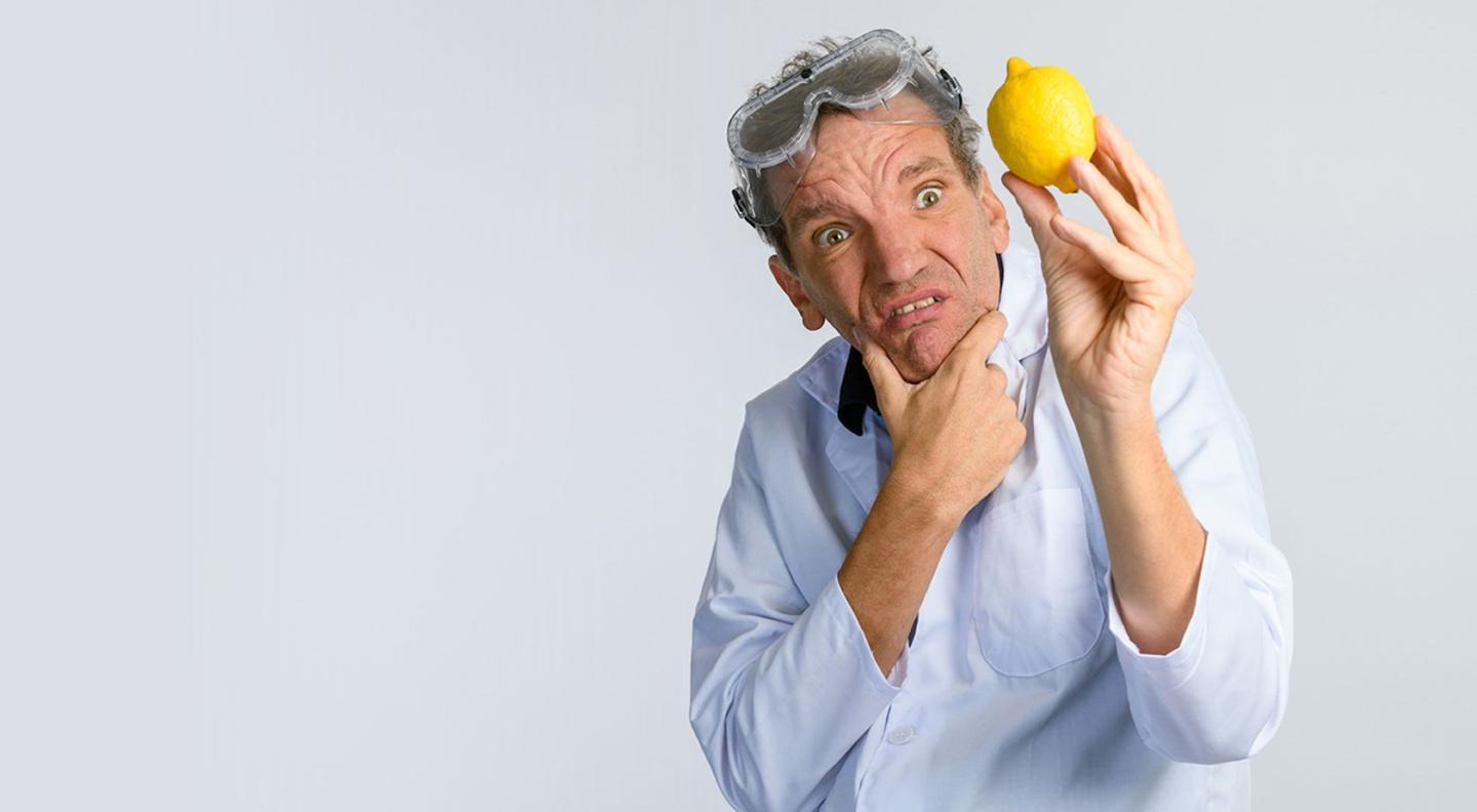 Henning Wehn wearing a lab coat with goggles on his head looking at a lemon he is holding.