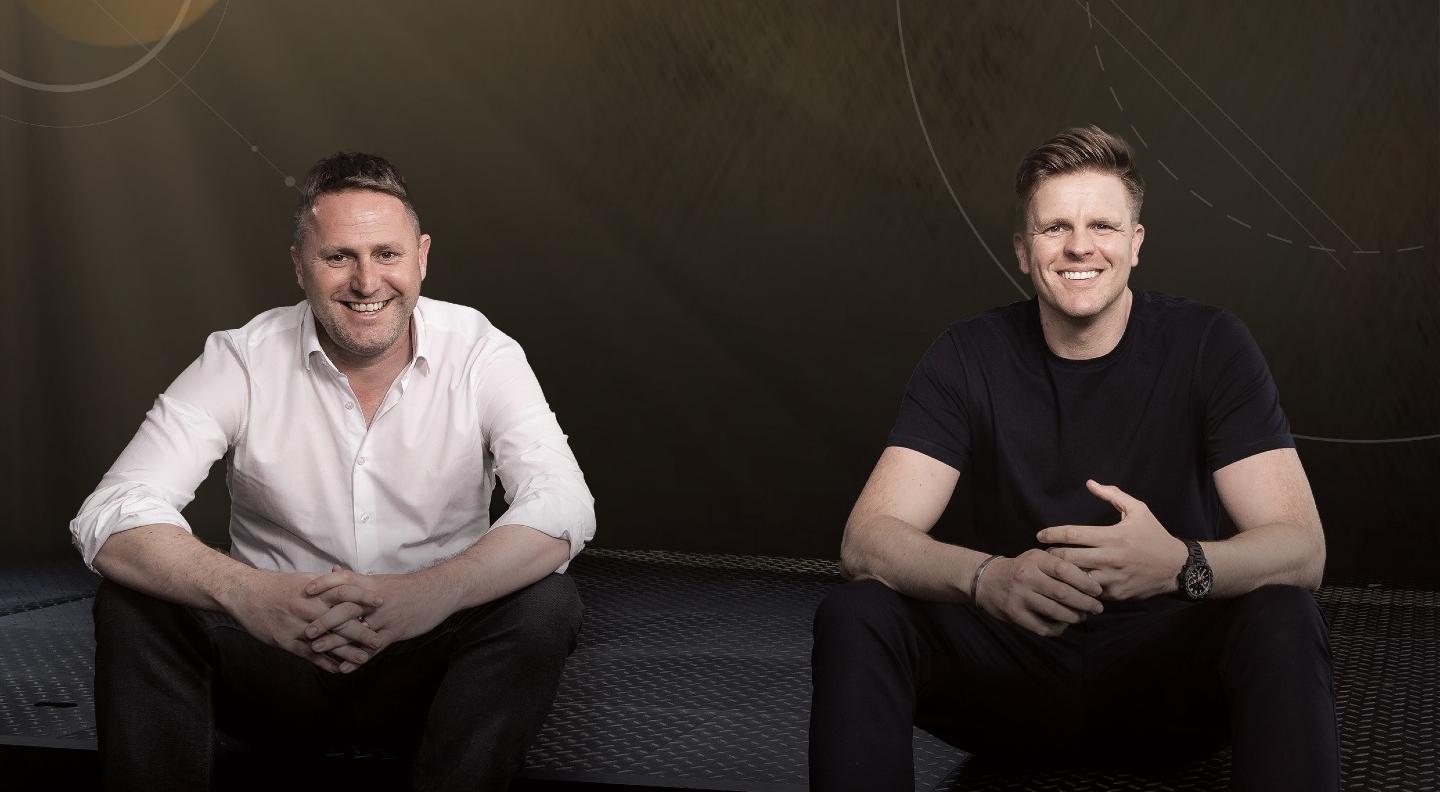 Jake Humphrey and Professor Damien Hughes sitting site by side against a dark background, resting their arms on their knees and smiling widely at the camera