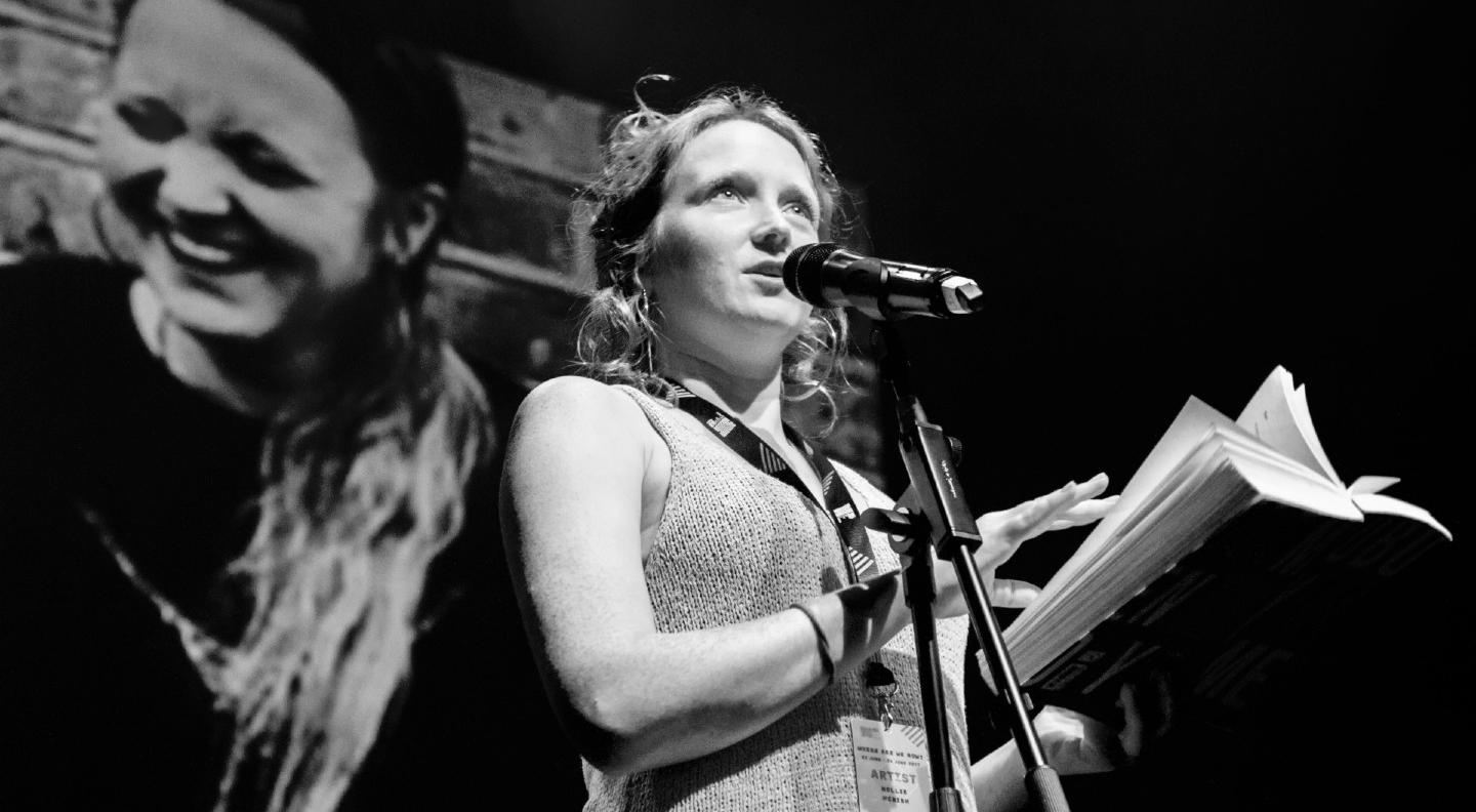 Image is black and white. Hollie McNish stands at a microphone with a book in her hand. There is an image of her laughing on the wall behind her
