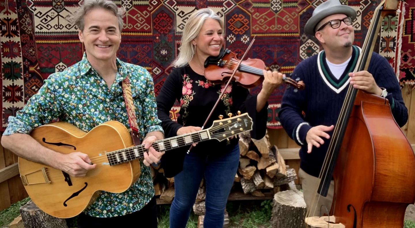 The three members of Hot Club of Cowtown - two men and one woman - are shown full length playing their instruments in an outside setting with grass under their feet, a patterned rug and logs of wood behind them. They all smile widely. From left to right they are playing guitar, violin and double bass
