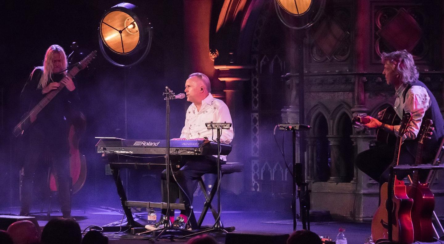 An image of Howard Jones and trio performing on stage in what looks like a church. Howard sits in the centre playing keyboard with two large arc lights directed at the audience on either side of him