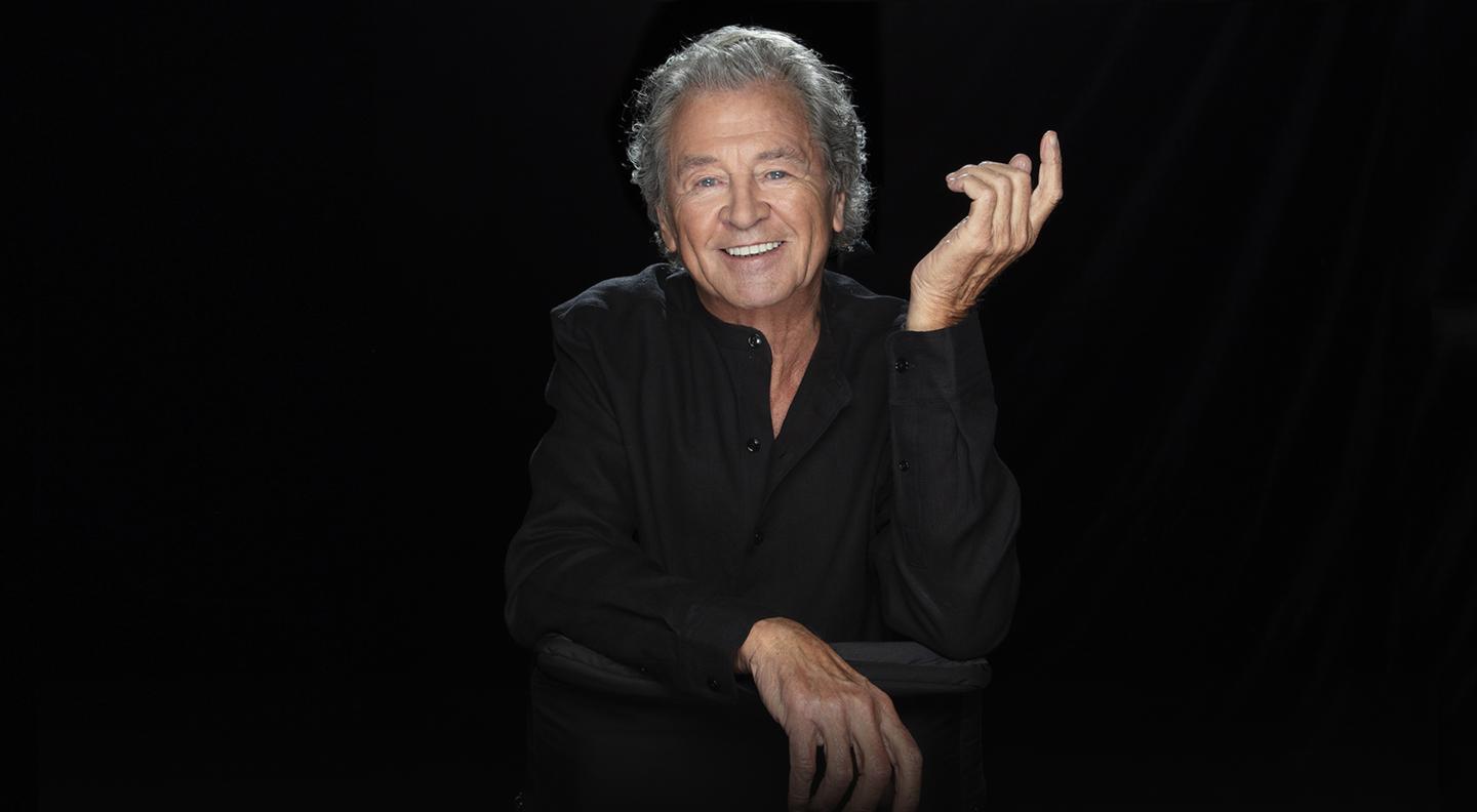 Ian Gillan standing with his hands clasped in front of him in front of a grey background