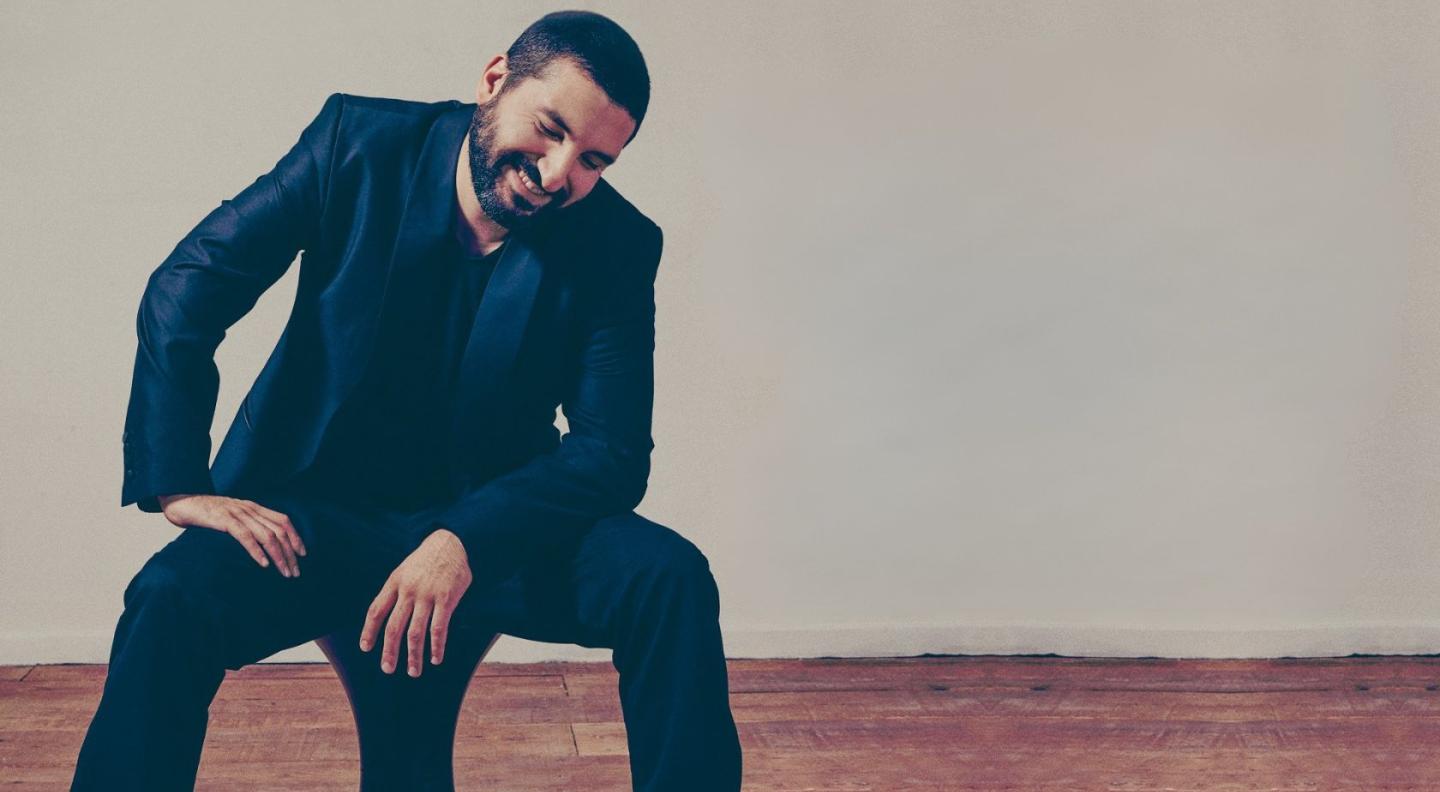 Ibrahim Maalouf sitting on a stool on a wooden floor wearing an all black suit