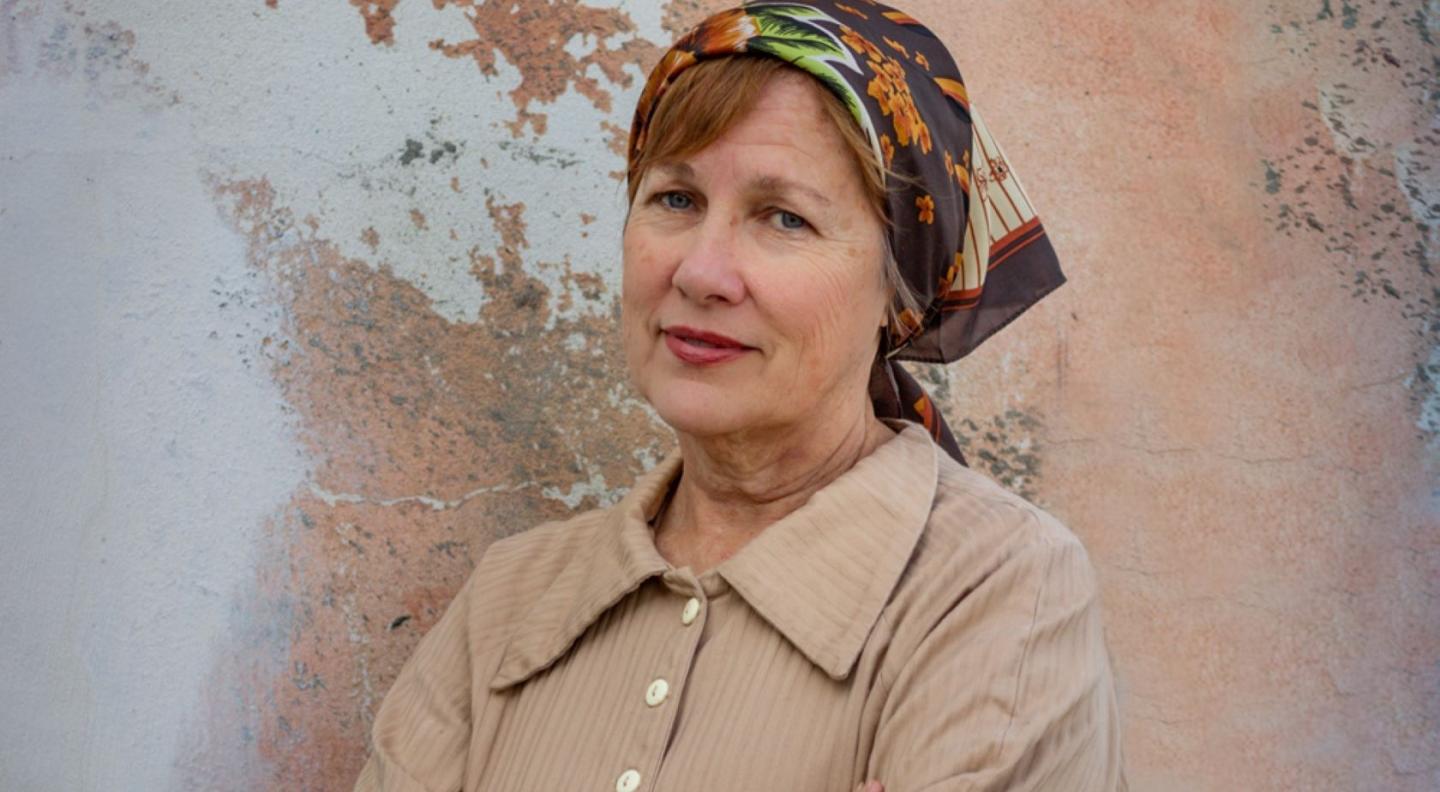 An older white woman with red hair wearing a patterned head scarf and beige shirt stands in front of a rough partly painted wall looking at the camera with her head on one side and her arms folded