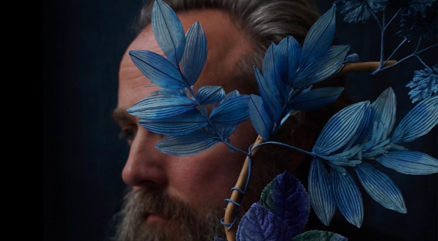 an image of a white man with a beard looking sideways, his face is obscured by blue leaves
