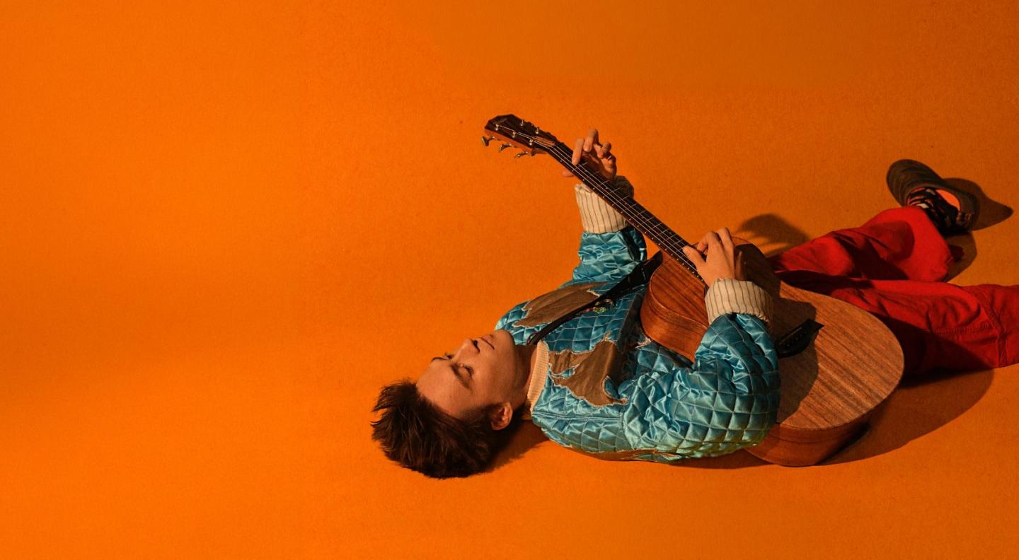 Jacob Collier lying on an orange backdrop, playing the guitar