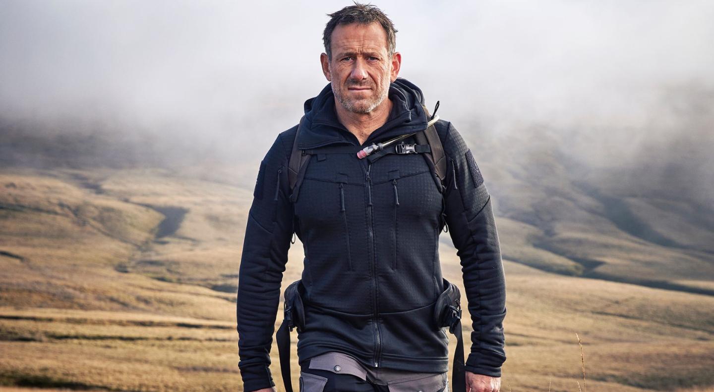 Jason Fox, a white man with short hair, walking towards the camera. Behind him is a hilly scene