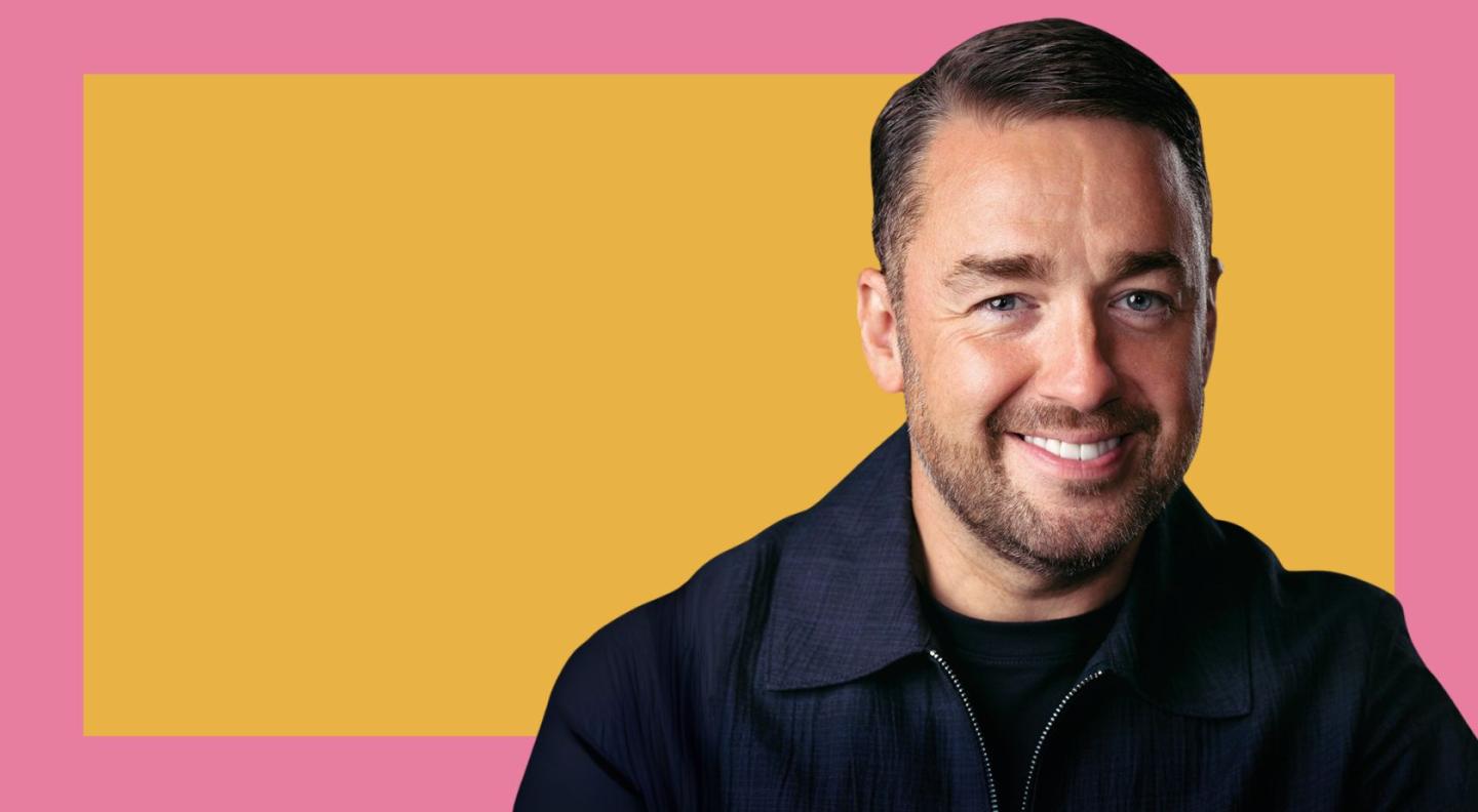 Jason Manford smiling in front of a yellow and pink background