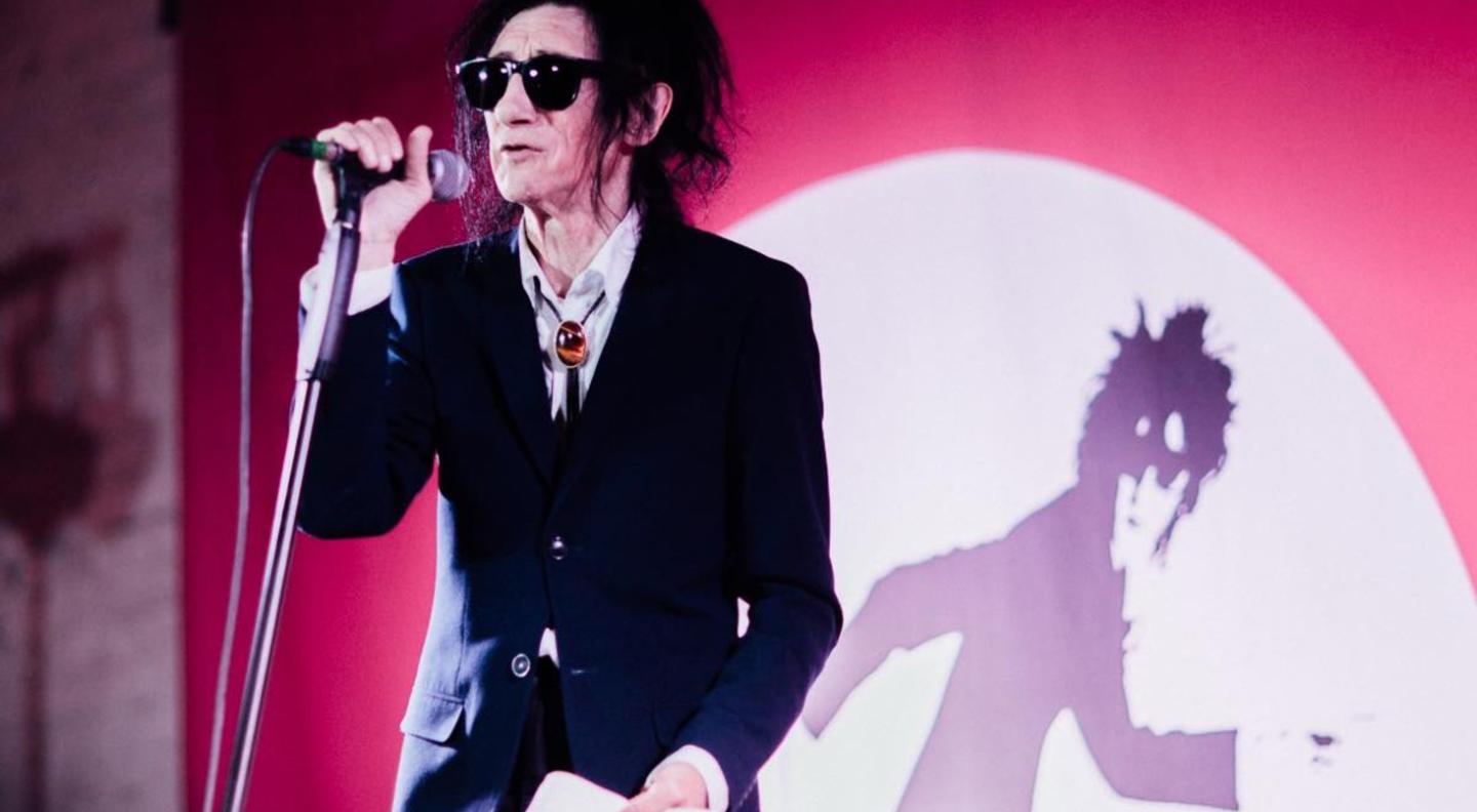 John Cooper Clark, wearing dark glasses, a dark jacket and tie and white shirt holds a microphone on a stand in his right hand and a book in his left hand. He is on stage in front of a red background with a white semi-circle containing an artistic impression of him in black and white.