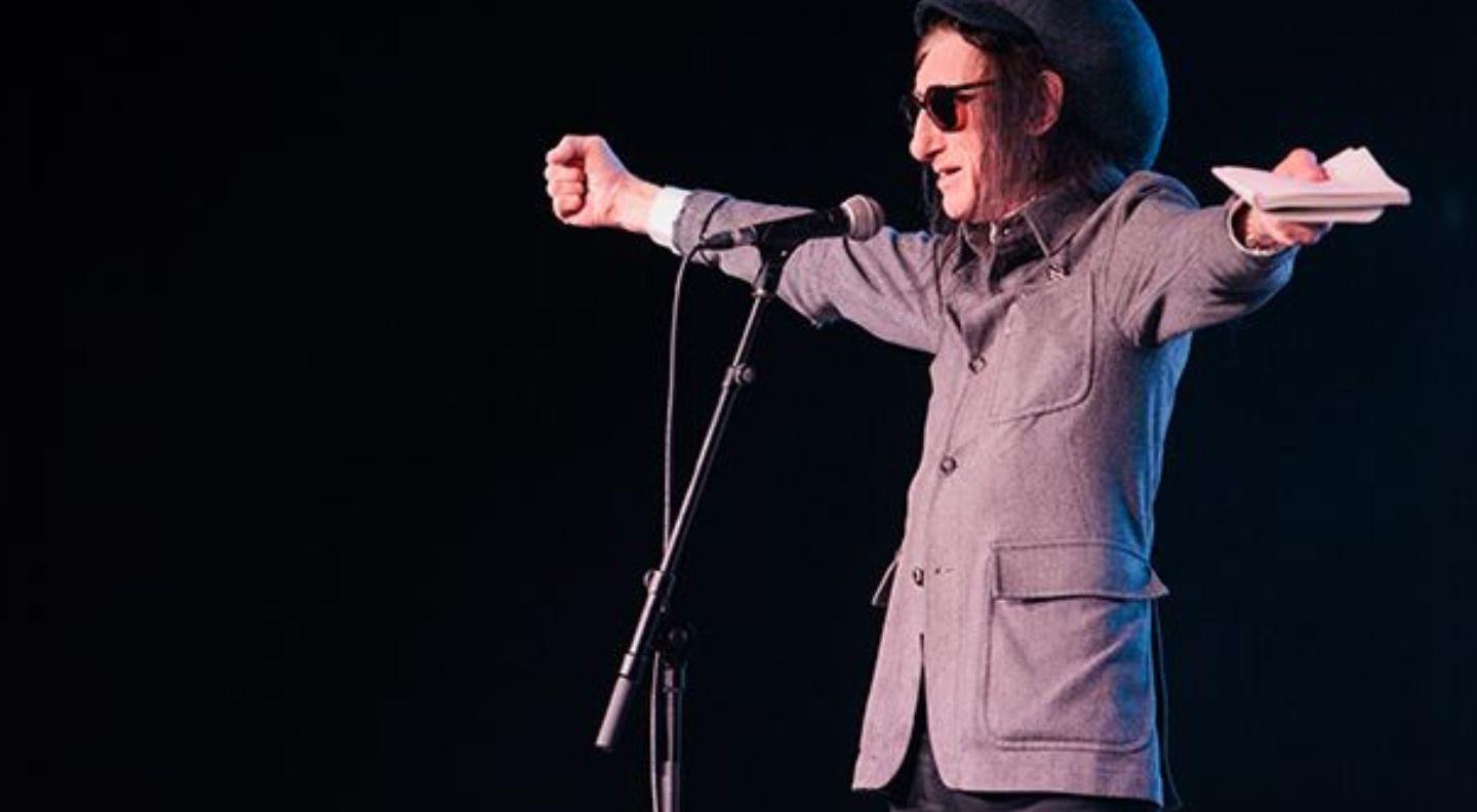 A white man with long dark hair tucked up into a big hat, wearing a grey jacket and sunglasses, speaks into a microphone on stage. 