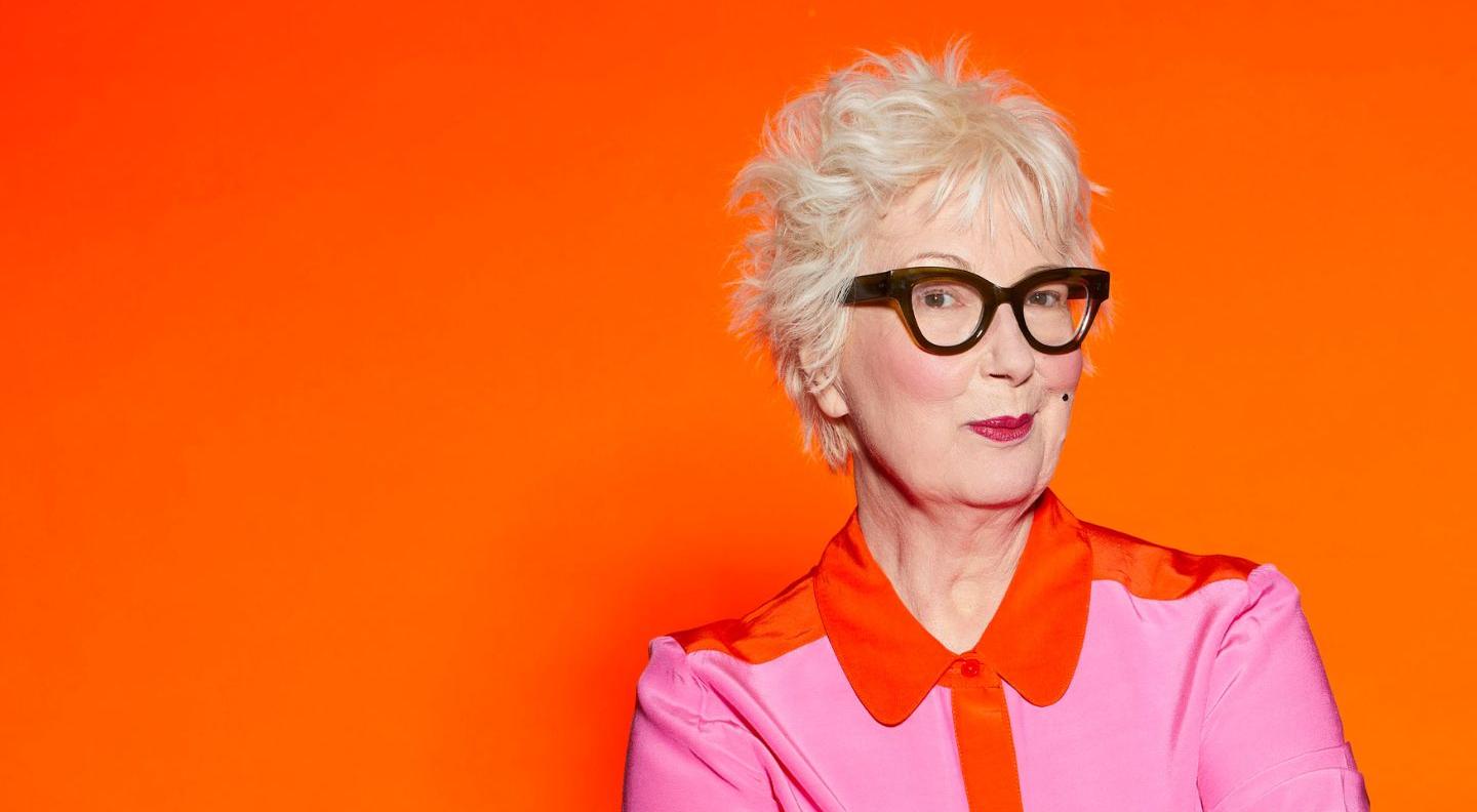 Jenny Eclair is a white woman with short, spiky white hair and dark framed glasses. She wears a bright pink shirt with an orange collar, and stands against an orange background