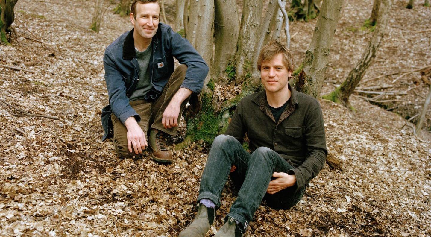 Johnny Flynn and Robert MacFarlane sit on leaf-covered ground, surrounded by trees. Both are white men with brown hair and wear neutral-toned clothes