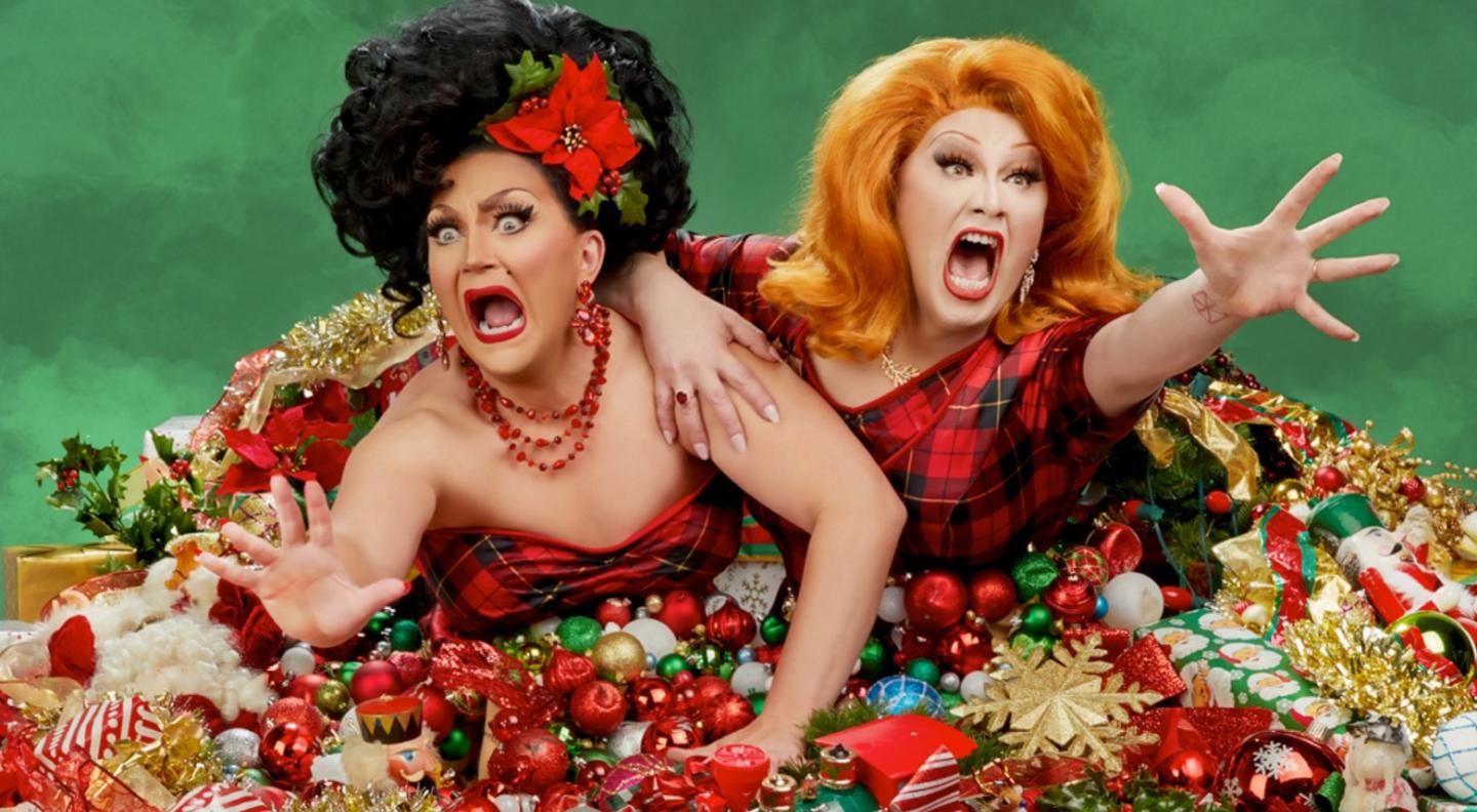 BenDeLaCreme and Jinkx Monsoon are seen from the waist up in full drag queen make-up. BenDeLaCreme has a bouffant black wig with a poinsettia flower in one side and large red dangly earrings. Jinkx Monsoon has a shoulder length ginger wig, manicured nails and a ring with a red stone. They are both dressed in tartan and reach out towards the camera from a mountain of baubles and Christmas decorations with a looks of horror on their faces.