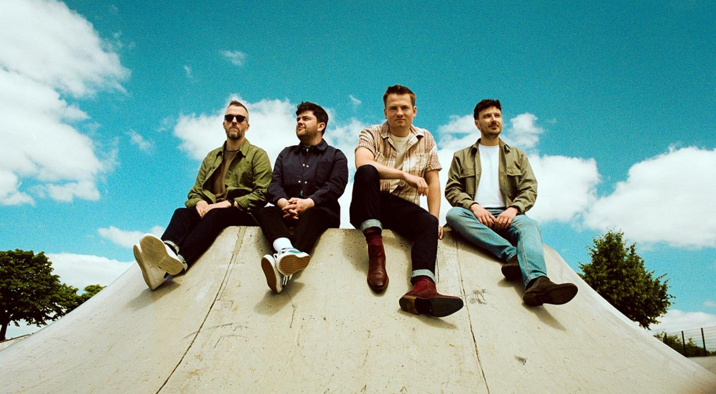 Tide Lines - four young white men - sit in casual clothing at the top of a skate ramp with a bright blue sky and white clouds behind them