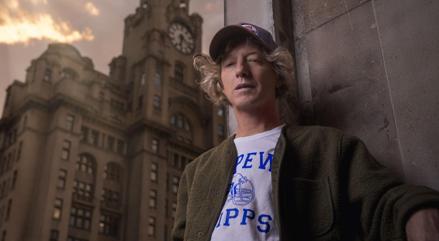 John Power, wearing a baseball cap, white t-shirt and green jacket leaning against a wall in front of an old building.