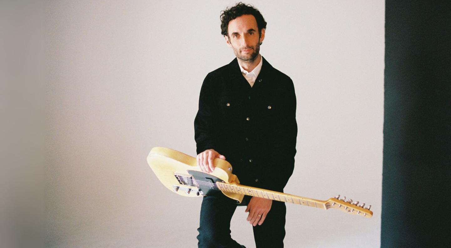 Julian Lege, holding a guitar in front of a white background