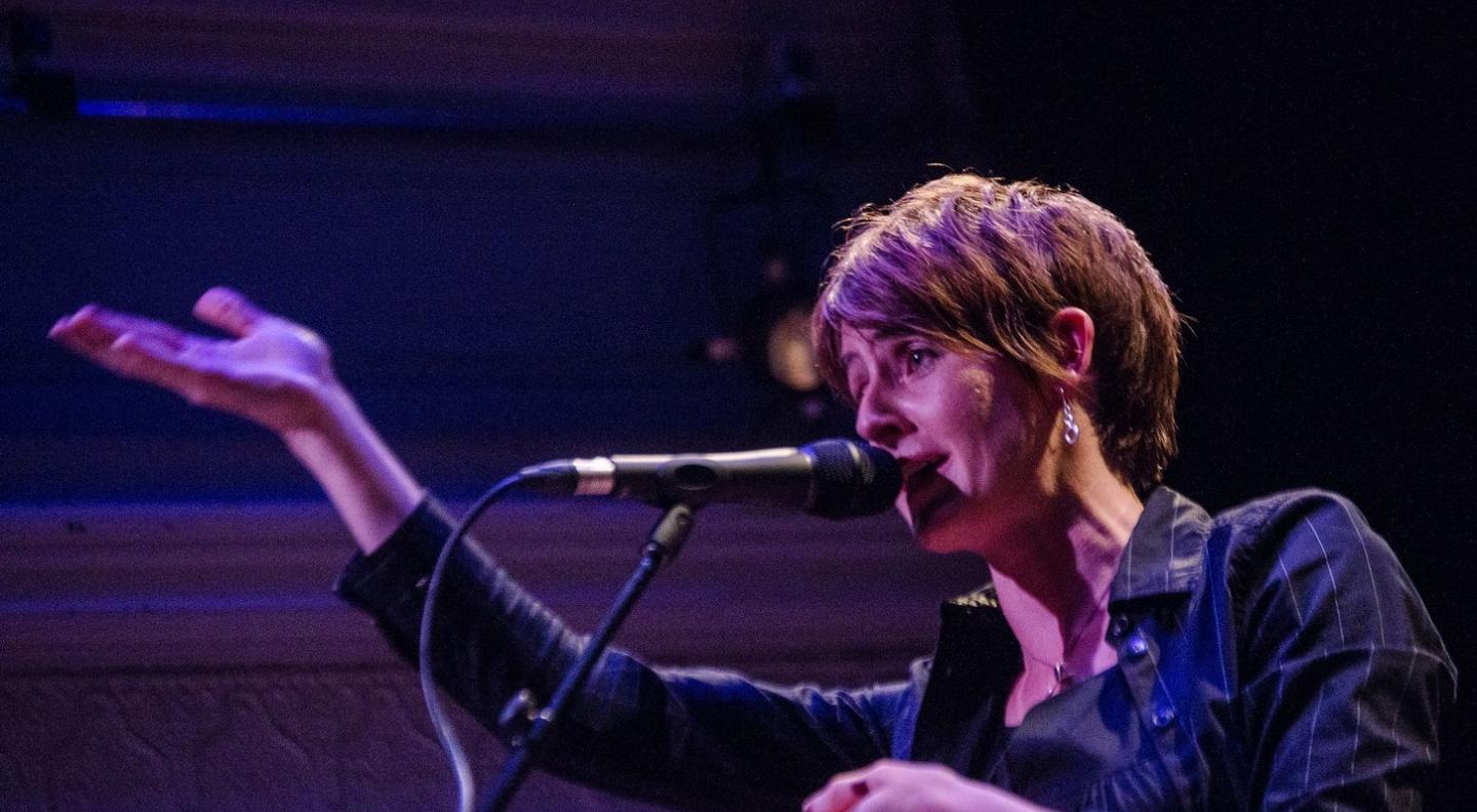 A white woman with short hair, Karine Polwart, faces to the left holding her arm out in front of her and singing into a microphone