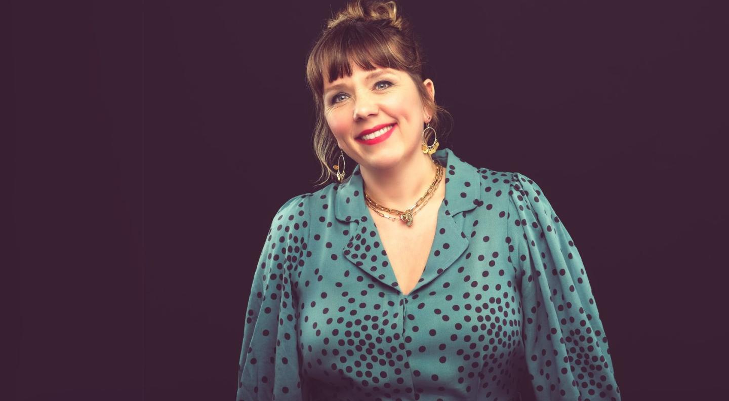 Kerry Godliman, a white woman in her fifties, standing in front of a black background, smiling at the camera. She is wearing a blue shirt with black spots.