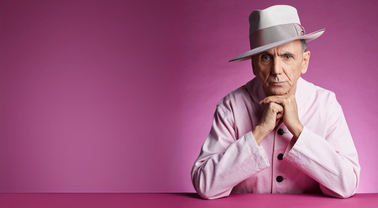 Kevin Rowland wearing a light pink jacket and hat, sitting in front of a pink background with his chin resting on his hands