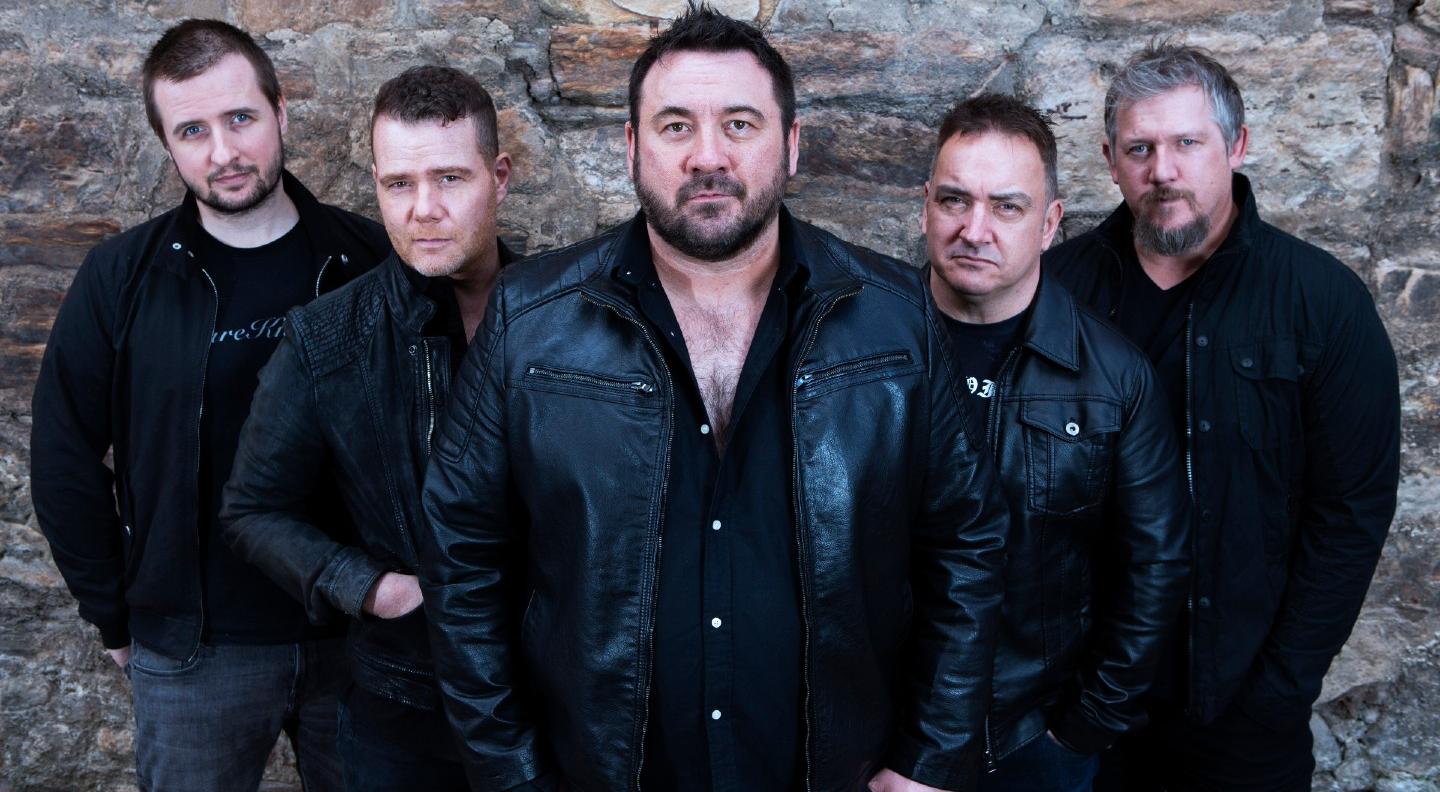 The five male members of King King, dressed in black stand in a line against a stone wall, all looking out at the camera. Photo taken by Graham Milne