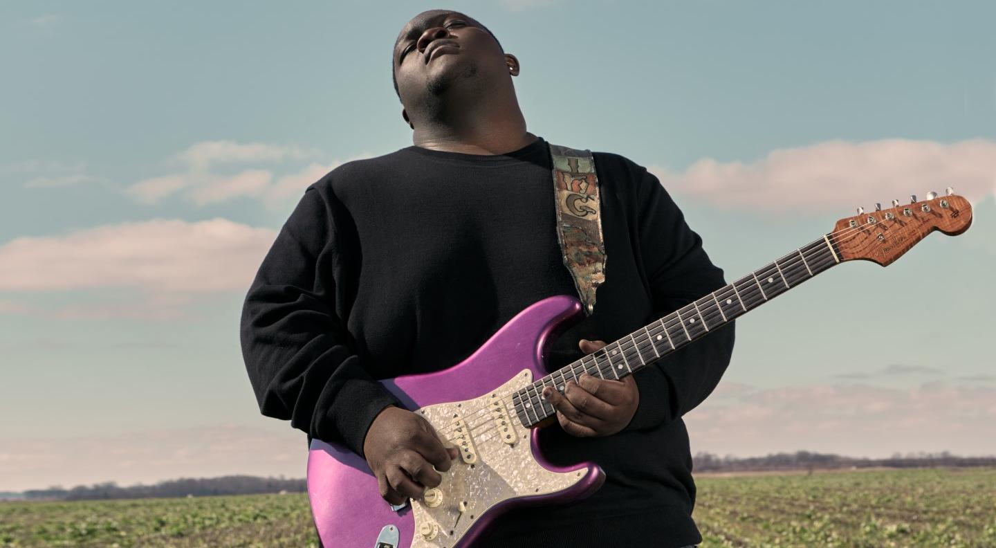Christone “Kingfish” Ingram plays a purple electric guitar in a green field