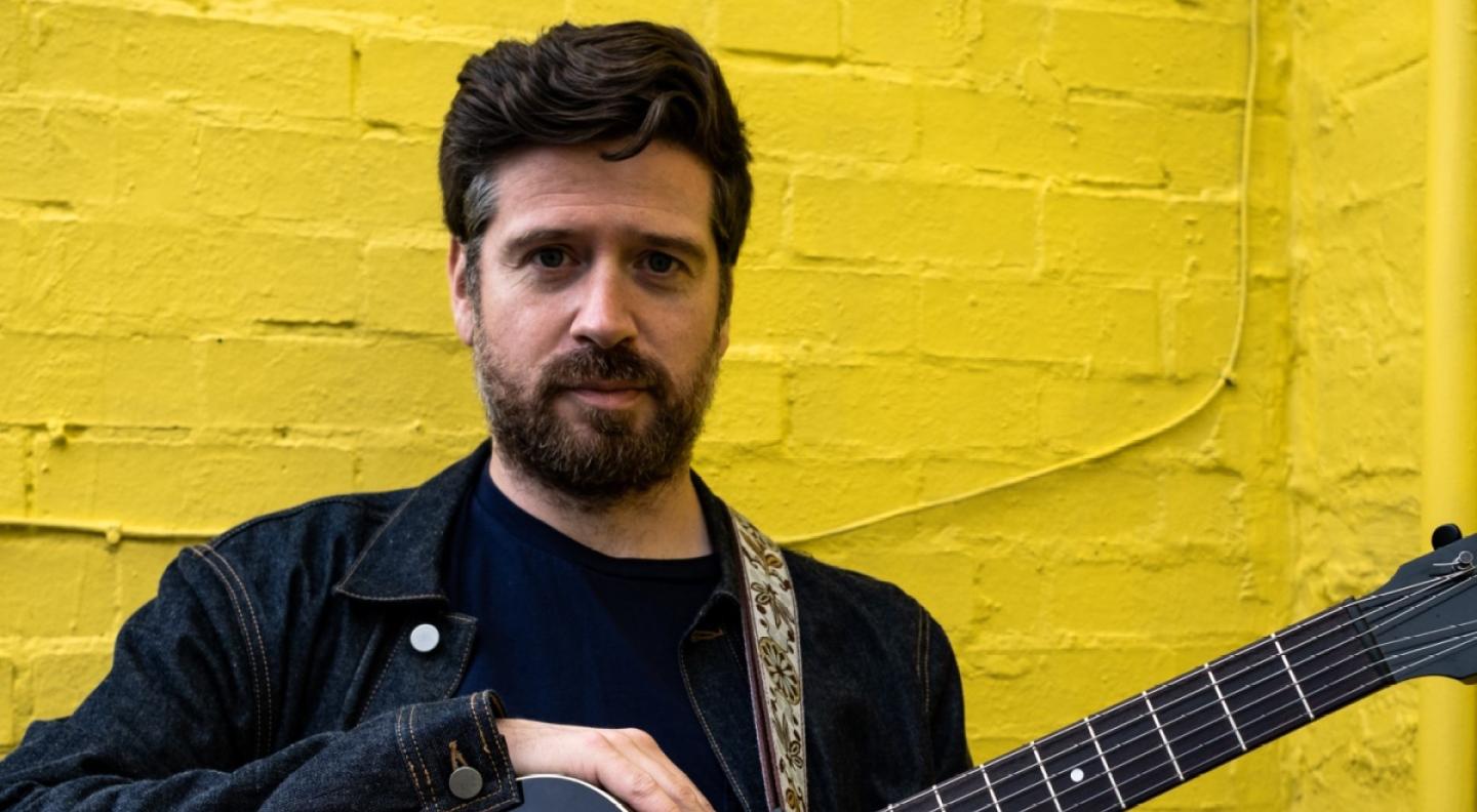 Kris Drever stands with a guitar against a yellow brick backdrop