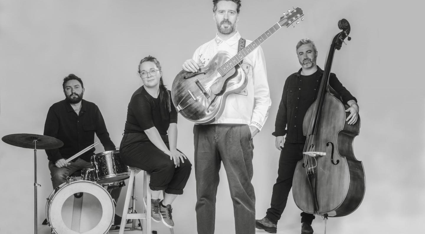 A black and white image of three white men and one white woman. One of the men stands in front of the others holding a guitar across his body. The other men stand holding a cello and sit behind a set of drums with the woman sitting on a high stool in between them