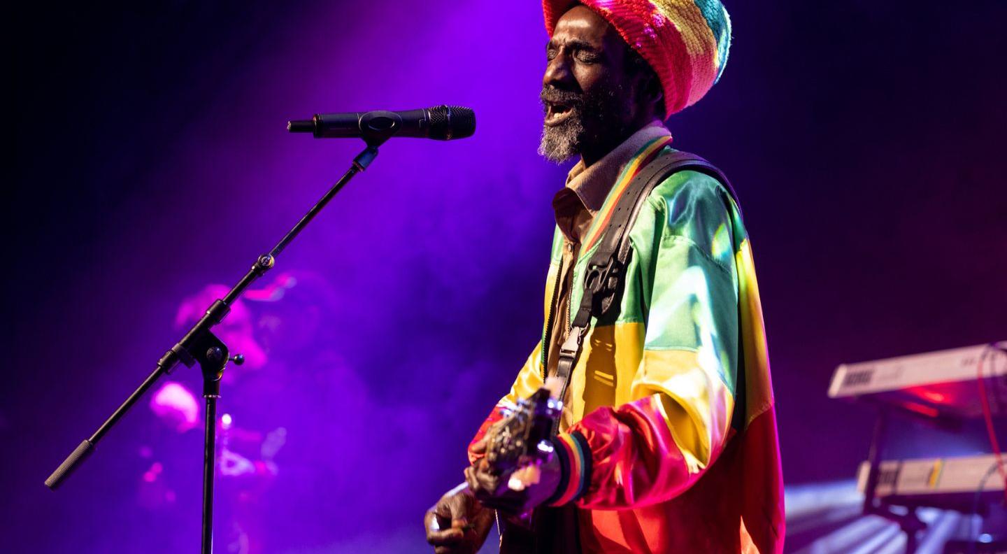 A black man sings into a microphone on stage while holding a guitar, wearing a green, yellow and red tracksuit jacket and large knitted hat in the same colours.