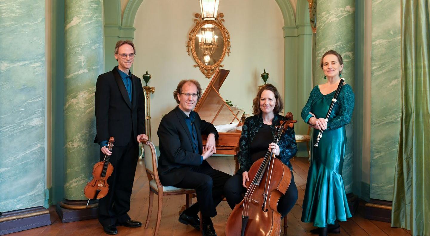 Four musicians stand in a green marble room. A man in a black suit holds a violin, and stands next to another man sat at the bench of a harpsichord. Next to them a woman sits holding her cello, and on the far right a woman in a green dress holds a dark wooden flute