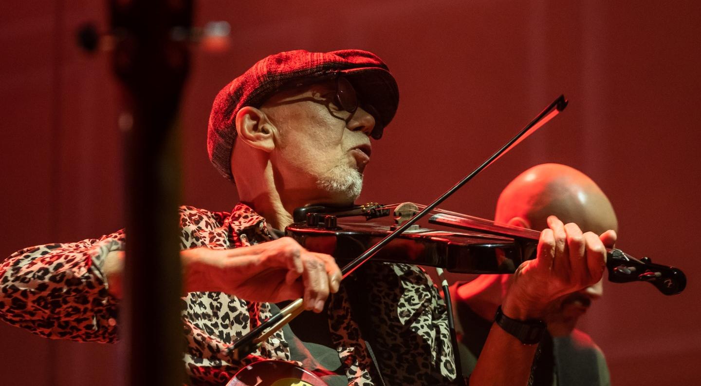 Rod Clements, in a flat cap and glasses, plays a fiddle, with a gitar also hanging around his shouders.