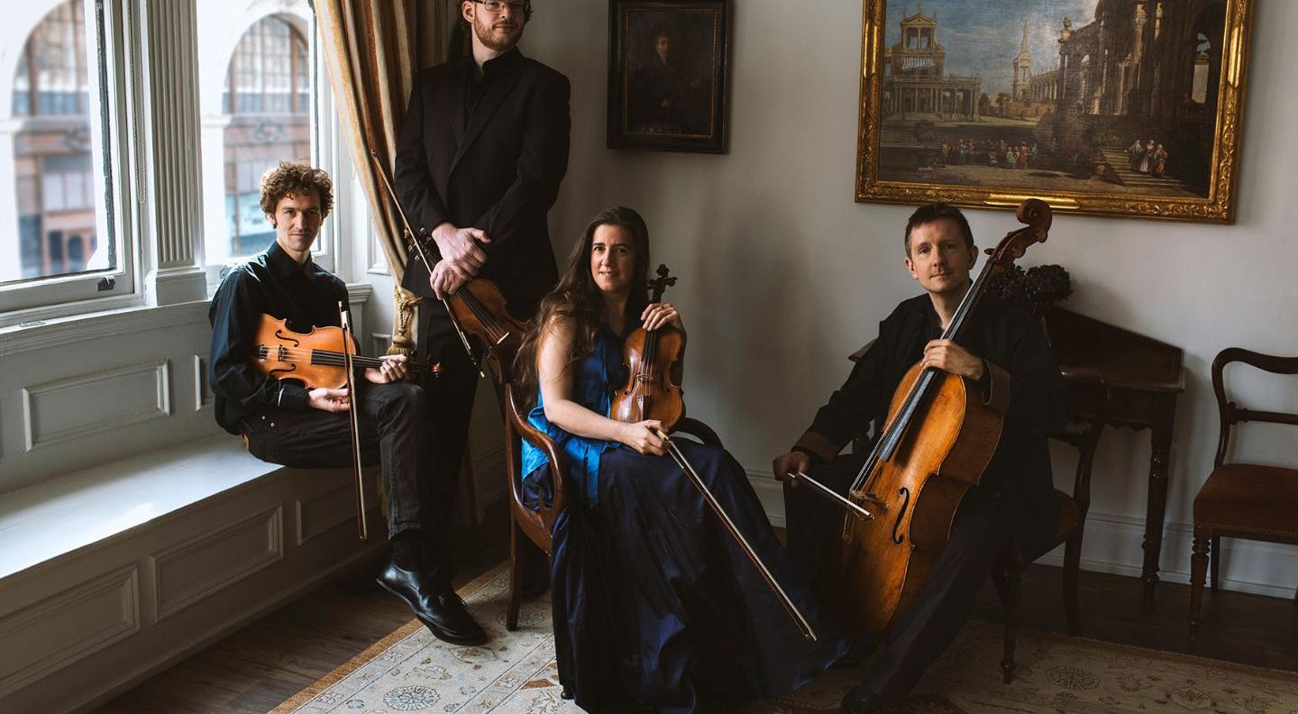 Four musicians sit in an ornately decorated Georgian room, holding their instruments. A man and a woman hold violins, one man has a viola, and one man has a cello 