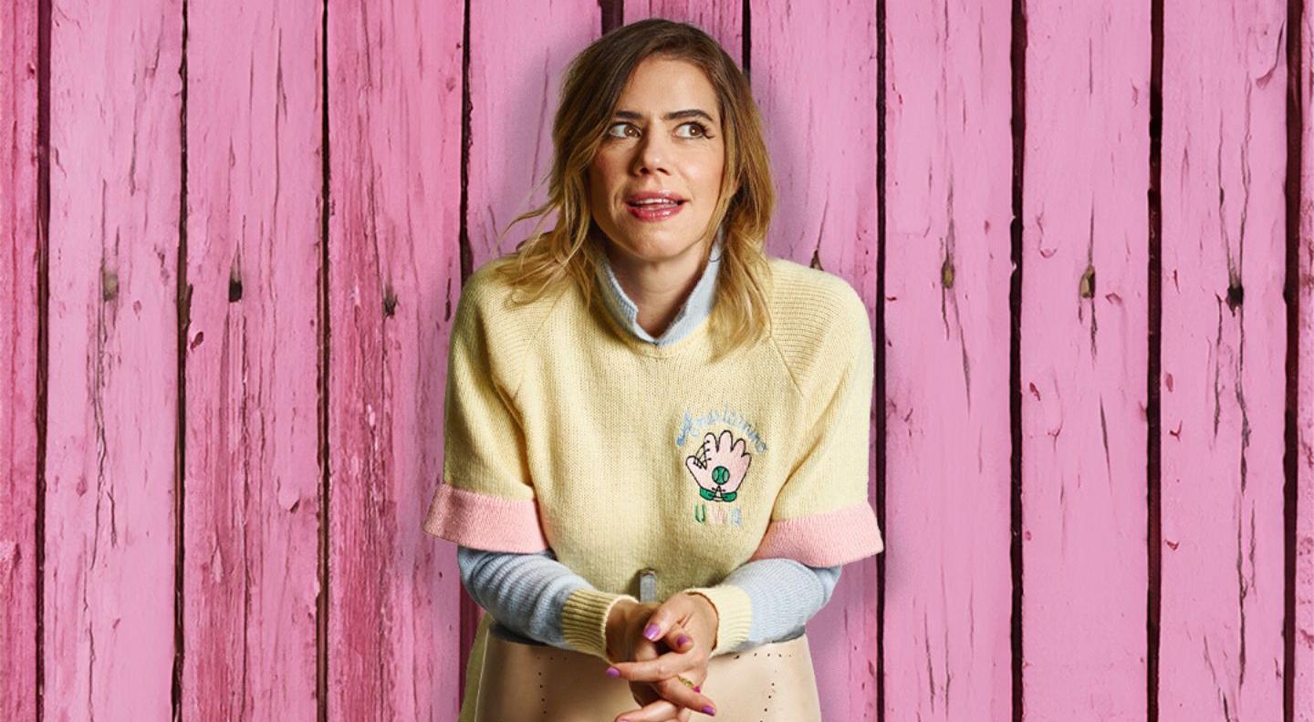 Lou Sanders, a white woman wearing a yellow t shirt, stands in front of a pink wooden background