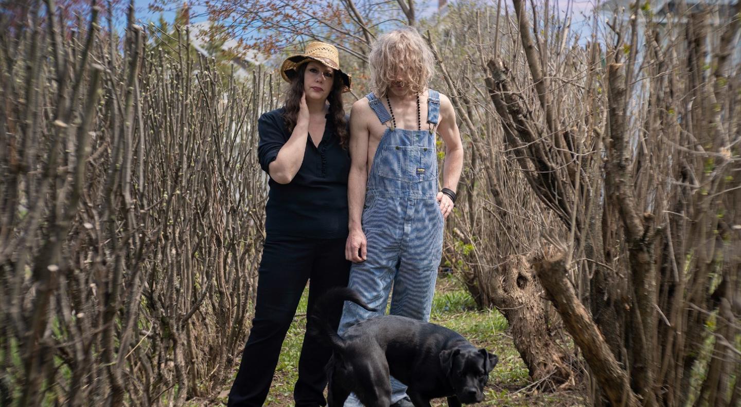 A full length shot of the two members of Low - Mimi Parker and Alan Sparhawk who stand in between two rows of leafless, pruned trees or vines. Hills or the sea is visible in the background and a black dog is in front of them. Mimi looks directly out at the camera and holds her right hand up to her face. Alan has his head bowed and his shoulder length blonde curly hair hides his face
