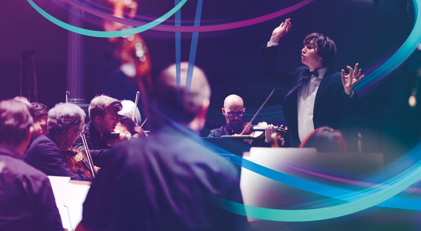 A young man with long hair conducts an orchestra, wearing a tuxedo