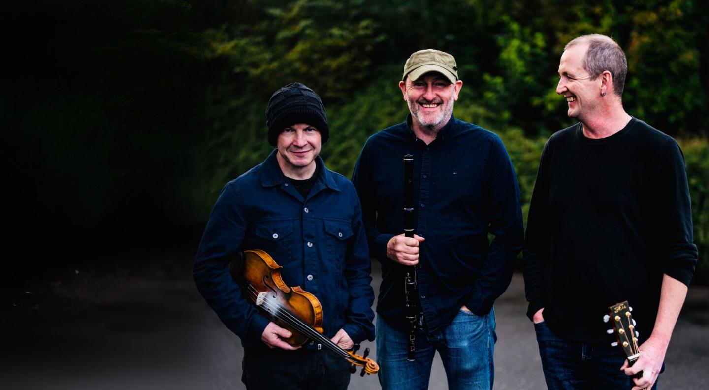 three white middle-aged men holding folk instruments with dark green bushes in the background