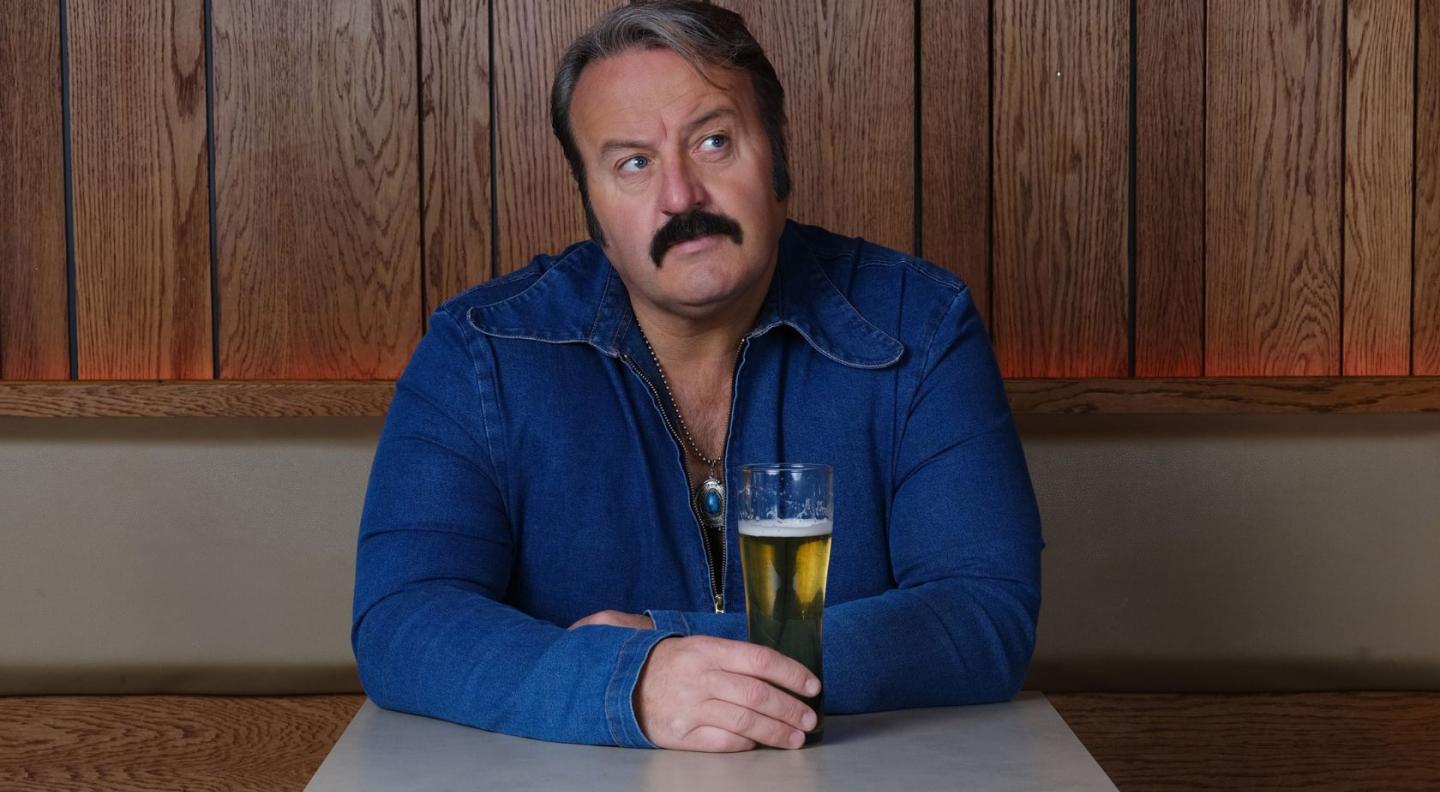 Mike Bubbins, wearing a denim jumpsuit, sitting at a table holding a pint glass full of beer