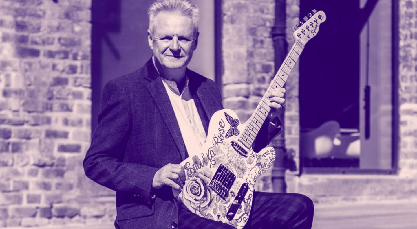 A blue-toned image of an older white man sitting on a pavement holding a guitar