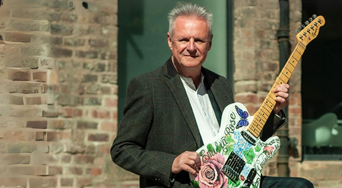 Mike McGeary - a white, middle-aged man with white hair, holds a green guitar in front of a building
