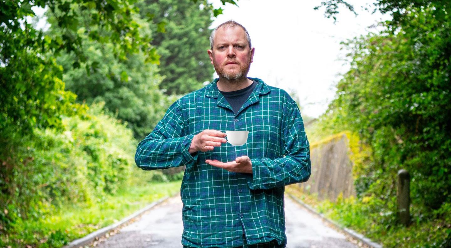 Miles Jupp, a white bald man, stands in a road lined by trees. He's wearing blue tartan pyjamas and holding a cup of tea