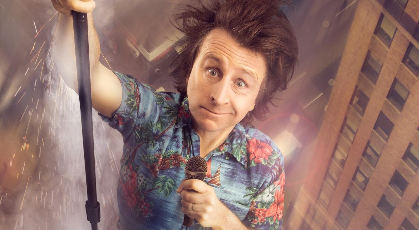 Milton Jones is holding on to a microphone stand looking like he's dangling above skyscrapers. He wears a flowered shirt, has messy brown hair and a slightly concerned look on his face