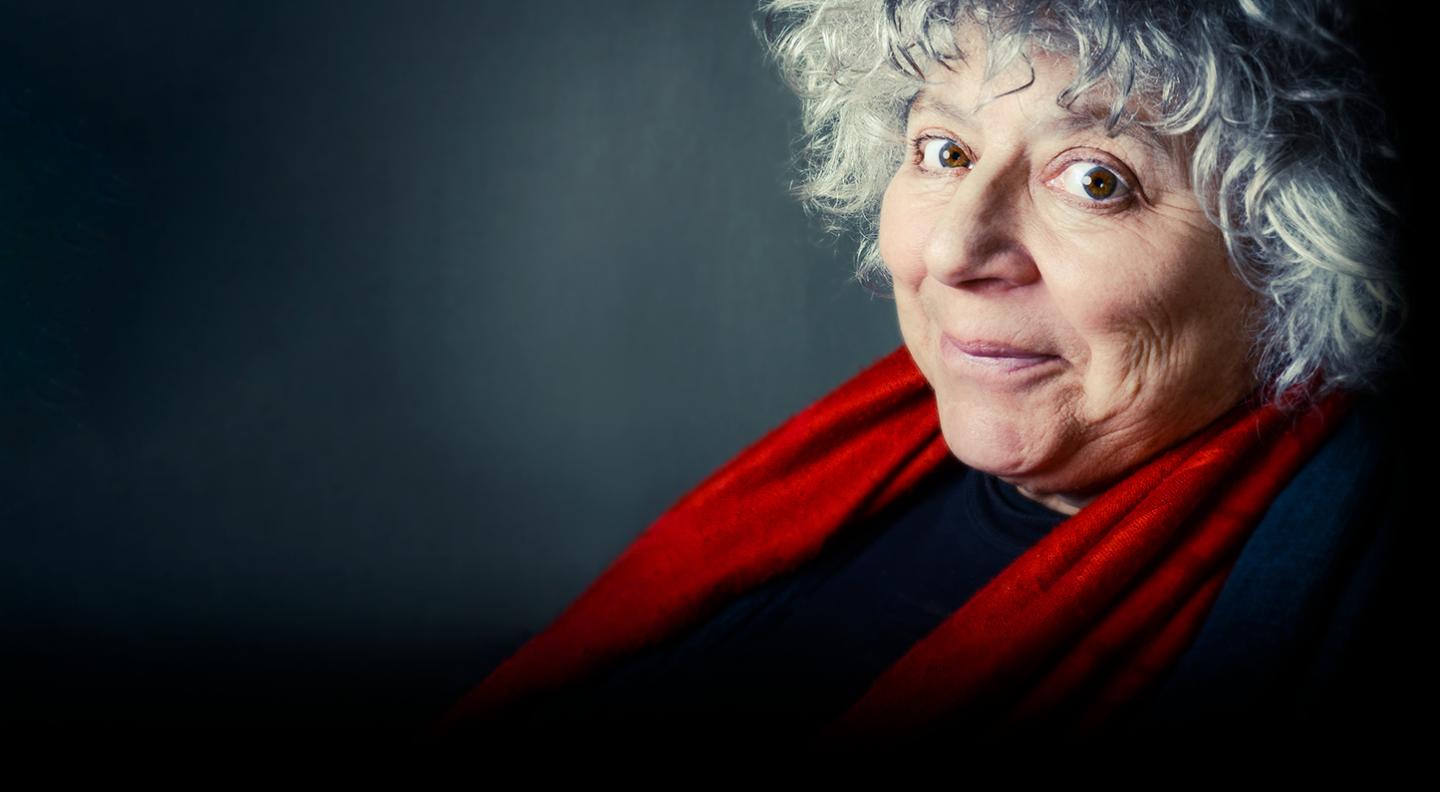A mid shot of Miriam Margolyes against a dark background. She wears a red scarf and looks out at the camera smiling
