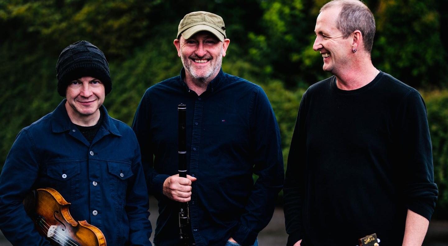 Three men wearing dark clothes laught together, standing against a background of trees